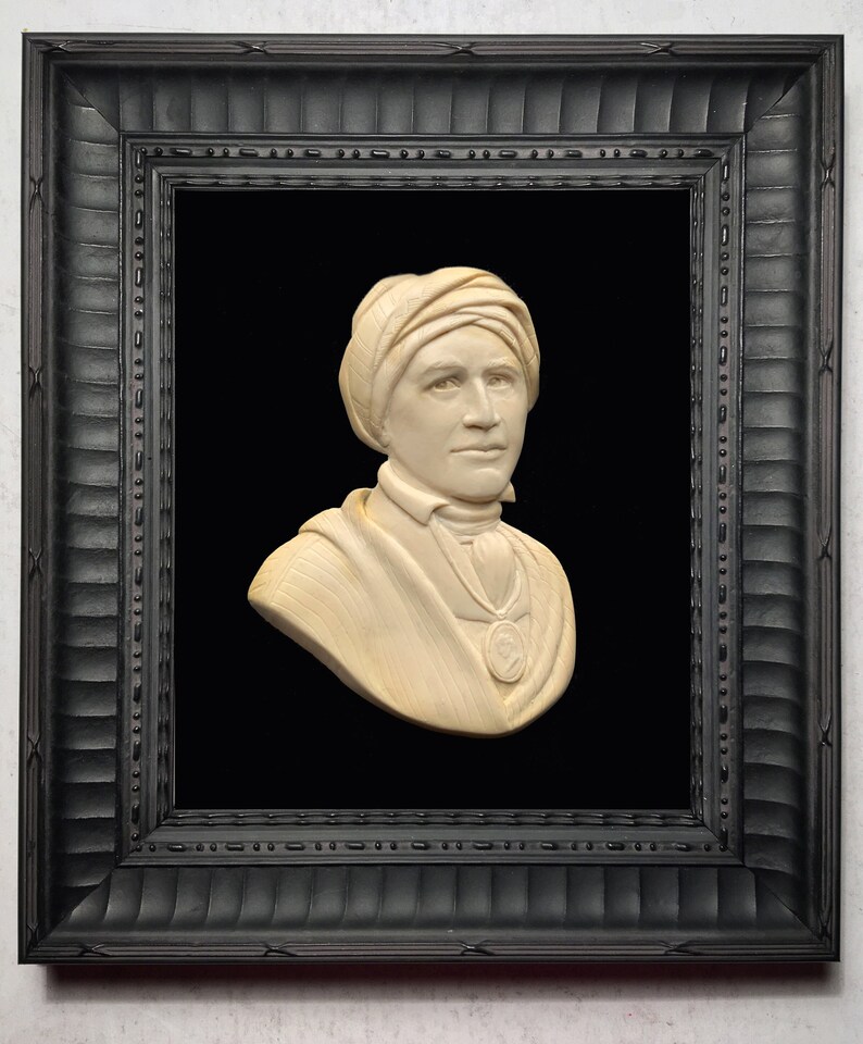 May include: A white ivory bust sculpture of a person wearing a turban, framed in a black wood frame with a black velvet mount.