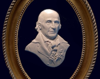 May include: A white ceramic bust of a man in a gold oval frame. The man is wearing a white shirt with a white cravat and a dark jacket. He has a white wig and a serious expression.