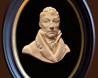 May include: A cream-coloured bust sculpture of a man in a framed oval. The man is wearing a dark jacket with a white collar and a white cravat. The frame is black with a gold trim.
