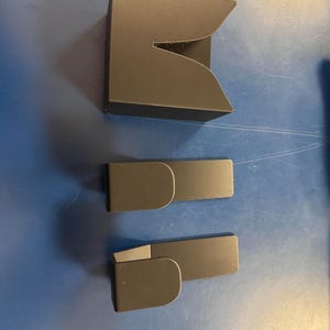 May include: Three black plastic wall hooks, two are rectangular and one is a curved shape. The hooks are designed to be mounted on a wall.