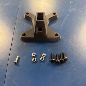 May include: A black plastic base with four screw holes and a square opening in the center. The base is accompanied by four silver nuts, three black screws, and one silver screw.