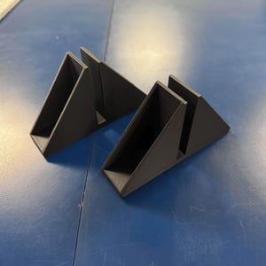 May include: Two black plastic triangular stands with a slot in the middle. The stands are on a blue surface.