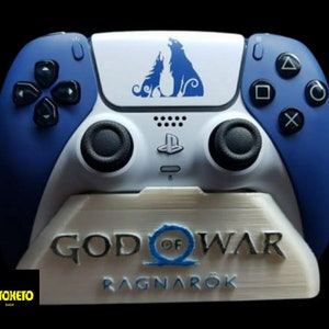 May include: A white and blue PlayStation 5 controller with a wolf design on the top. The controller is sitting on a white stand with the words "God of War Ragnarök" printed on it.