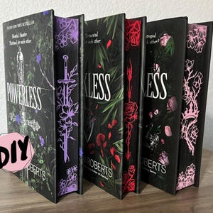 Könnte beinhalten: Eine Sammlung von fünf Büchern mit schwarzen Einbänden. Die Buchrücken zeigen Titel wie "POWERLESS" und "RECKLESS" in Weiß, mit floralen und Schwertmotiven in Lila und Rot. Die Bücher sind auf einer Holzoberfläche ausgestellt.