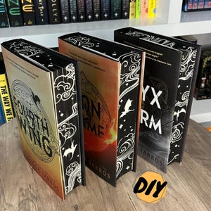 May include: Four hardcover books with decorative black and white edges are displayed on a wooden surface. The book covers feature titles like "Fourth Wing" and "Onyx Storm." The books are part of a series, with a DIY sticker in the foreground.