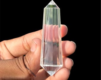 Könnte beinhalten: Ein klarer Quarz-Kristall-Obelisk mit facettiertem Design. Der Kristall wird vor einem schwarzen Hintergrund gehalten, der seine Transparenz und geometrische Form zeigt. Der Kristall ist etwa 13 cm hoch.