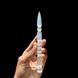 K&ouml;nnte beinhalten: Ein klarer, l&auml;nglicher Kristallstab mit einer Spitze und einem strukturierten Griff. Der Stab wird vor einem schwarzen Hintergrund gehalten. Der Kristallstab ist etwa 18 cm lang.