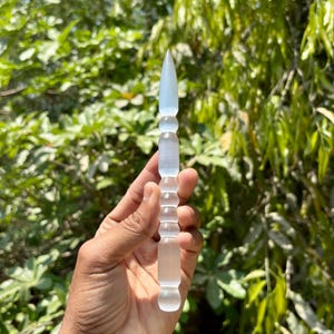 K&ouml;nnte beinhalten: Ein klarer, l&auml;nglicher Kristallstab mit einer Spitze und einem zylindrischen Griff. Der Stab wird vor einem Hintergrund aus gr&uuml;nem Laub gehalten, was seine durchscheinende Qualit&auml;t und sein detailliertes Design hervorhebt. Der Stab ist etwa 15 cm lang.