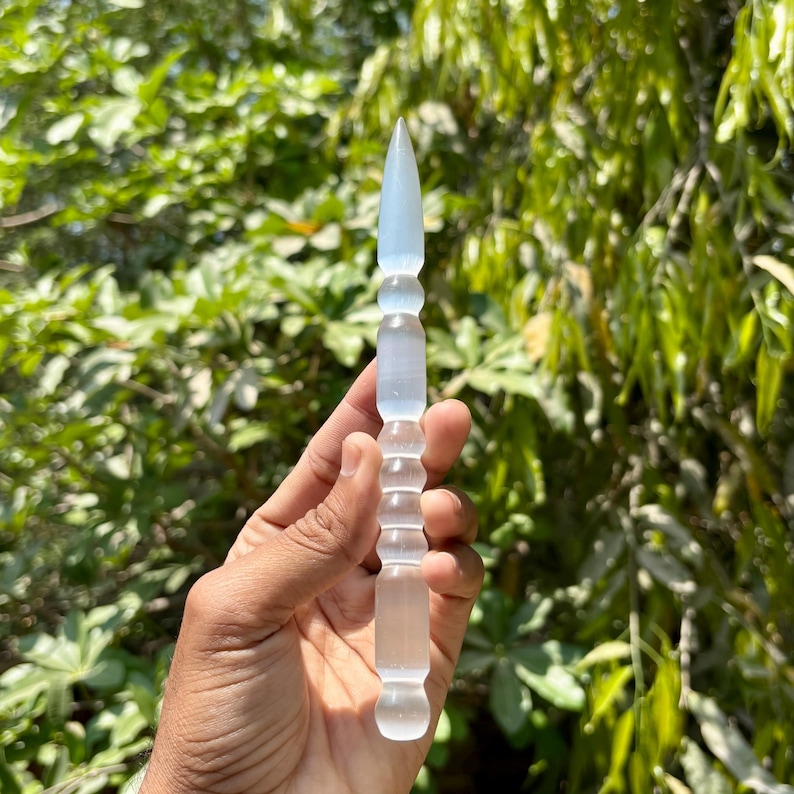 K&ouml;nnte beinhalten: Ein klarer, l&auml;nglicher Kristallstab mit einer Spitze und einem zylindrischen Griff. Der Stab wird vor einem Hintergrund aus gr&uuml;nem Laub gehalten, was seine durchscheinende Qualit&auml;t und sein detailliertes Design hervorhebt. Der Stab ist etwa 15 cm lang.