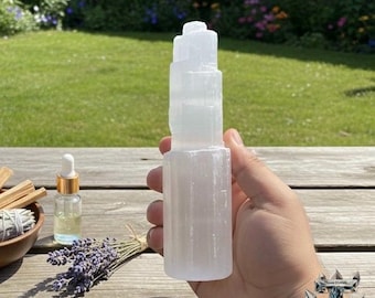 Könnte beinhalten: Eine klare, geschichtete Selenitturm wird in einer Hand gehalten. Eine kleine Glasflasche mit goldenem Verschluss und ein Lavendelstrauß liegen auf einem Holztisch. Eine Schale mit Salbei ist ebenfalls zu sehen.