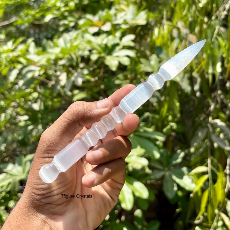 K&ouml;nnte beinhalten: Ein klarer, l&auml;nglicher Selenitstab mit einer Spitze und einem geriffelten Griff. Der Kristallstab wird vor einem Hintergrund aus gr&uuml;nem Laub gehalten. Der Stab ist etwa 15 cm lang.