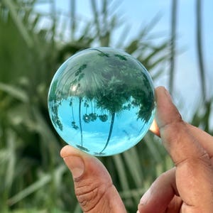 Può includere: Una sfera di vetro trasparente che riflette un cielo blu e palme. La palla è tenuta in una mano, con gli alberi e il cielo che appaiono capovolti all'interno. L'immagine crea una rappresentazione artistica unica della natura.