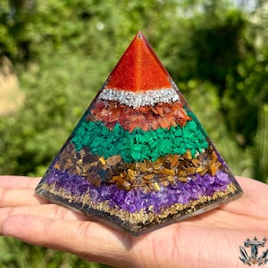 May include: A layered pyramid-shaped decorative object with a red, silver, green, brown, and purple design. The pyramid is held in a hand, with a blurred green background.
