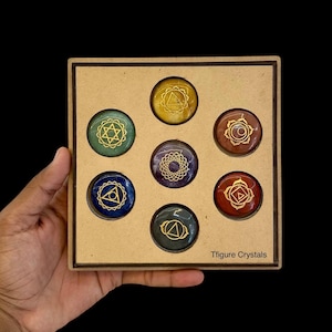 Op de afbeelding: Een set van zeven chakra stenen in een houten doos. Elke steen heeft een andere kleur, waaronder geel, groen, blauw, paars, bruin en rood. Elke steen heeft een goudkleurig chakra symbool. De doos wordt door een hand vastgehouden. De tekst "Tfigure Crystals" is zichtbaar.