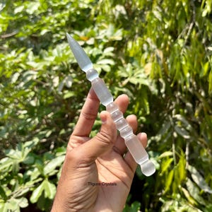 K&ouml;nnte beinhalten: Ein klarer Kristallstab mit einer Spitze und einer Reihe von abgerundeten und zylindrischen Segmenten. Der Stab wird vor einem Hintergrund aus gr&uuml;nem Laub gehalten. Der Text "Tfigure Crystals" ist sichtbar.