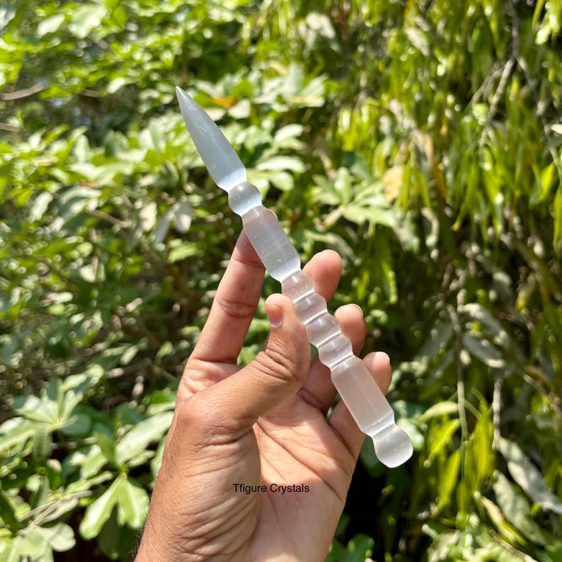 K&ouml;nnte beinhalten: Ein klarer Kristallstab mit einer Spitze und einer Reihe von abgerundeten und zylindrischen Segmenten. Der Stab wird vor einem Hintergrund aus gr&uuml;nem Laub gehalten. Der Text "Tfigure Crystals" ist sichtbar.