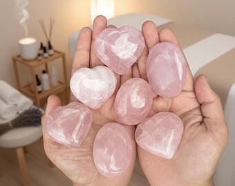 Puede incluir: Primer plano de manos sosteniendo varias piedras de cuarzo rosa pulidas. Las piedras tienen forma de corazón y ovalada, con un delicado color rosa. El fondo presenta una pequeña mesa de madera con suministros de aromaterapia.