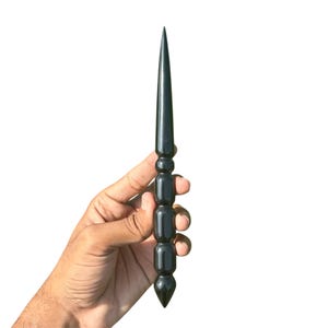 May include: A black, pointed wand with a decorative, rounded handle. The wand is held against a white background. The wand's tip is sharp and conical, and the handle has a series of stacked, cylindrical shapes.