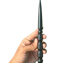 May include: A black, pointed wand with a decorative, rounded handle. The wand is held against a white background. The wand's tip is sharp and conical, and the handle has a series of stacked, cylindrical shapes.