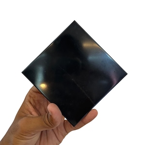 Può includere: Un oggetto quadrato nero lucido è tenuto in mano su sfondo bianco. L'oggetto ha una superficie riflettente, con una leggera lucentezza. I bordi sono affilati e ben definiti, suggerendo un design moderno.