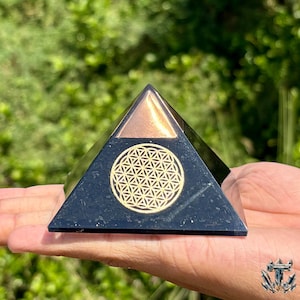 May include: A black pyramid with a copper-colored top and a gold-colored geometric design on the front. The pyramid is held in a hand against a blurred green background.