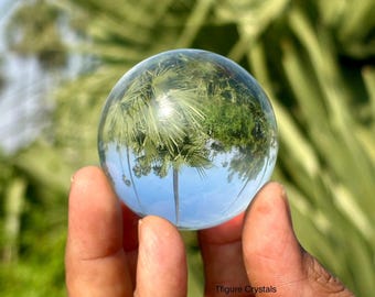 May include: A clear crystal ball reflects a palm tree and blue sky. The sphere is held in a hand, with a blurred green background of foliage. The text "Tfigure Crystals" is visible.