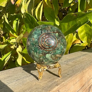 May include: A green crystal sphere with a copper spiral design, displayed on a small, ornate gold-colored stand. The sphere is composed of various shades of green and brown. The background features green foliage.