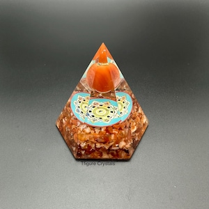 May include: A pyramid-shaped orgone crystal with a clear resin exterior. The pyramid contains orange-brown stones, a round orange stone, and a turquoise and gold design. The text "Tfigure Crystals" is at the base.