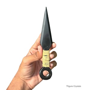 May include: A black kunai knife with a triangular blade and a ring at the base of the handle. The handle is wrapped with beige twine. The knife is held against a white background.
