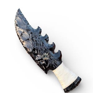May include: A black obsidian knife with a serrated edge and a white wrapped handle. The blade has a glossy, reflective surface. The handle is wrapped with white cord. The knife is displayed against a white background.