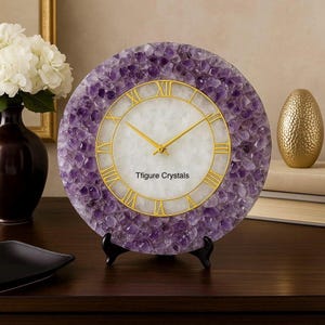 May include: Round amethyst crystal clock with gold-colored Roman numerals and hands. The clock face is white with the text "Tfigure Crystals". The clock is displayed on a black stand.