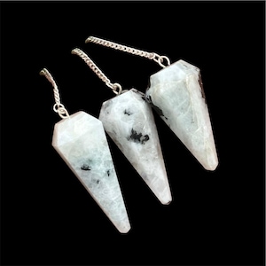 May include: Three white and gray crystal pendulums with silver chains. Each pendulum has a pointed, faceted shape. The crystals have black inclusions and are displayed against a black background.