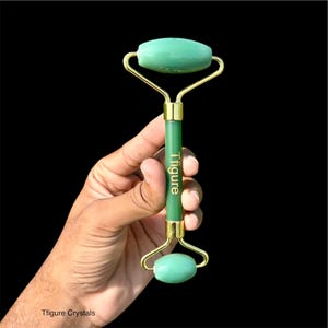 Op de afbeelding: Een groene jade gezichtsroller met goudkleurige metalen accenten. De roller heeft een handvat met de tekst "Tfigure" in goud gedrukt. De roller wordt tegen een zwarte achtergrond gehouden.