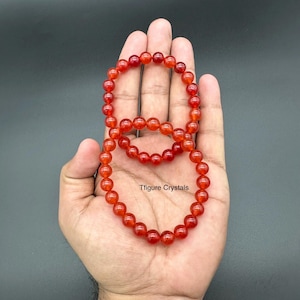 May include: Two strands of red-orange gemstone bead bracelets are displayed. The beads are round and appear to be approximately 0.3 inches in diameter. The bracelets are resting on a hand, with the text "Tfigure Crystals" visible.