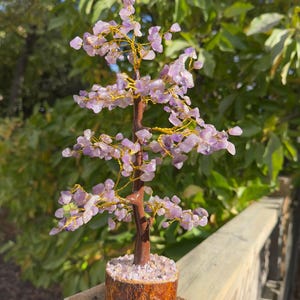 Op de afbeelding: Een decoratieve amethist boomsculptuur. De boom heeft een bruine stam, gouden draad takken en clusters van paarse amethist stenen. Het staat in een houten voet gevuld met kleine amethist chips. De achtergrond toont groen gebladerte.
