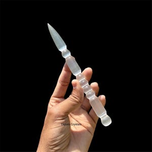 Puede incluir: Una varita de cristal transparente, con una punta afilada y un mango texturizado. La varita se sostiene sobre un fondo negro. La varita mide aproximadamente 18 cm de largo.