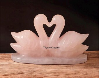 May include: A pink rose quartz sculpture of two swans forming a heart shape. The swans are on a base, and the text "Tfigure Crystals" is visible. The sculpture is displayed on a wooden surface.