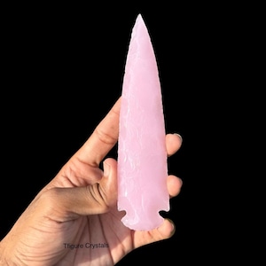 Puede incluir: Una punta de flecha de cristal rosa con punta y base dentada. La punta de flecha se sostiene en una mano sobre un fondo negro. El cristal tiene una superficie texturizada.