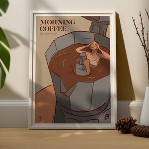 May include: A framed art print featuring a woman relaxing in a Moka pot filled with coffee. The print has the words "MORNING COFFEE" and "THE MOKA POT" in brown text. The artwork is in a white frame.