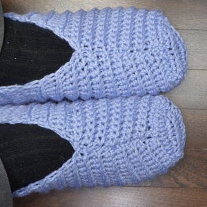 May include: A pair of light blue crocheted slippers with a ribbed design. The slippers are worn over black socks, with the legs of the person wearing them visible. The background is a dark brown wooden floor.