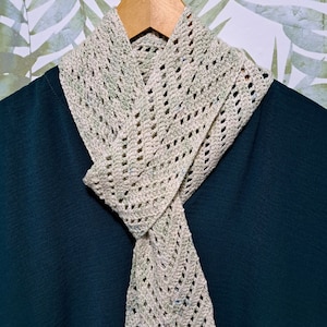 Peut inclure: Une écharpe en crochet beige avec un motif ajouré délicat, suspendue à un cintre en bois sur un fond de feuilles vertes.