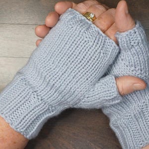 May include: A pair of light gray knitted fingerless gloves. The gloves are made of a ribbed knit fabric and have a thumb opening. The gloves are designed to keep hands warm while leaving fingers free. The gloves are a neutral color.