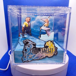 Peut inclure: Boîte d'exposition en acrylique transparent avec un diorama Final Fantasy X. La scène représente deux personnages sur un fond d'eau bleu avec le logo du jeu. La boîte est sur une base rotative blanche.
