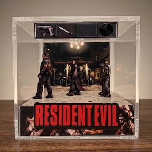 May include: A clear acrylic display case featuring miniature figures from the Resident Evil video game series. The case includes a pistol, knife, and a vinyl record. The Resident Evil logo is prominently displayed at the base.