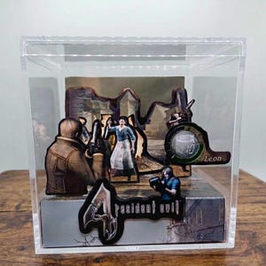 May include: A Resident Evil 4 diorama in a clear acrylic display case. The scene features character cutouts, including Leon, and a backdrop of a village. The Resident Evil 4 logo is visible on the base.