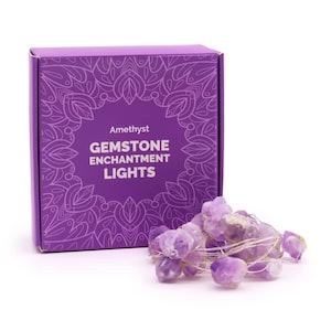 May include: A purple box with the words "Amethyst Gemstone Enchantment Lights" next to a string of amethyst gemstone lights. The box has a decorative floral design. The gemstones are a light purple colour.