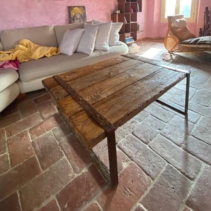 Könnte beinhalten: Ein rustikaler Holz-Couchtisch mit Metallbeinen steht in einem Wohnzimmer mit einer pinkfarbenen Wand und einem großen Gemälde einer Person in roter Kleidung. Der Raum hat einen Terrakotta-Fliesenboden.