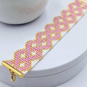 May include: A beaded bracelet featuring a repeating pink and white pattern. The bracelet is secured with a gold-colored clasp. The beads are small and create a textured surface. The bracelet is a stylish accessory.