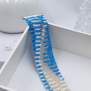 May include: A beaded bracelet with a pattern of blue and white beads. The bracelet has a silver clasp and is displayed on a white surface. The beads are small and create a textured surface.