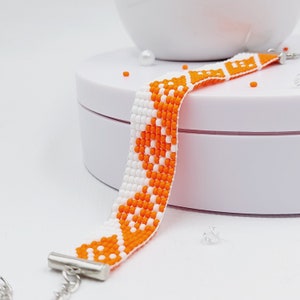 May include: A beaded bracelet featuring a white and orange geometric pattern. The bracelet is made of small, colorful beads and has a silver clasp. The design incorporates a repeating pattern of orange shapes against a white background.
